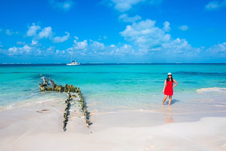 Kate Storm in a red dress standing on Playa Norte in Isla Mujeres. The remains of a dock are to her left.